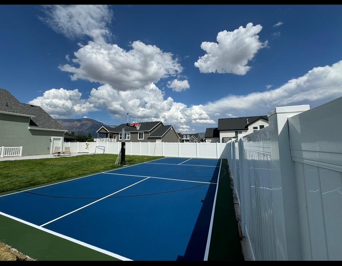 Blue pickleball court with green apron and black basketball lines in a white-fenced backyard with mountains behind
