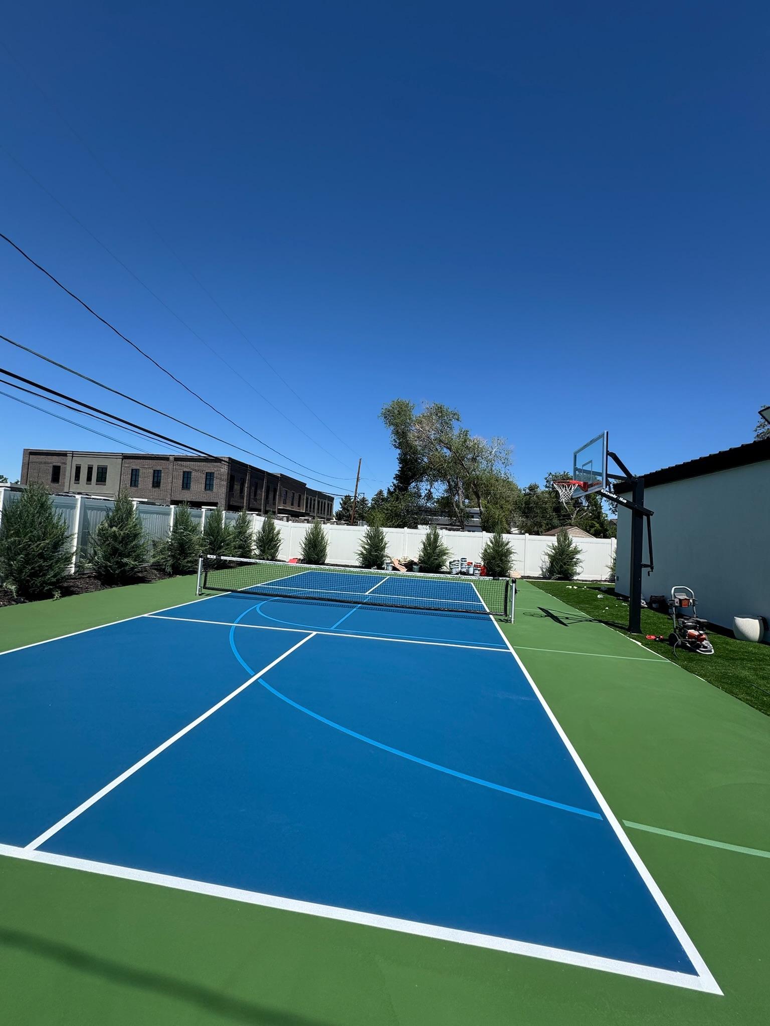 Two-tone blue pickleball court with green apron, net, and basketball hoop in a fenced backyard
