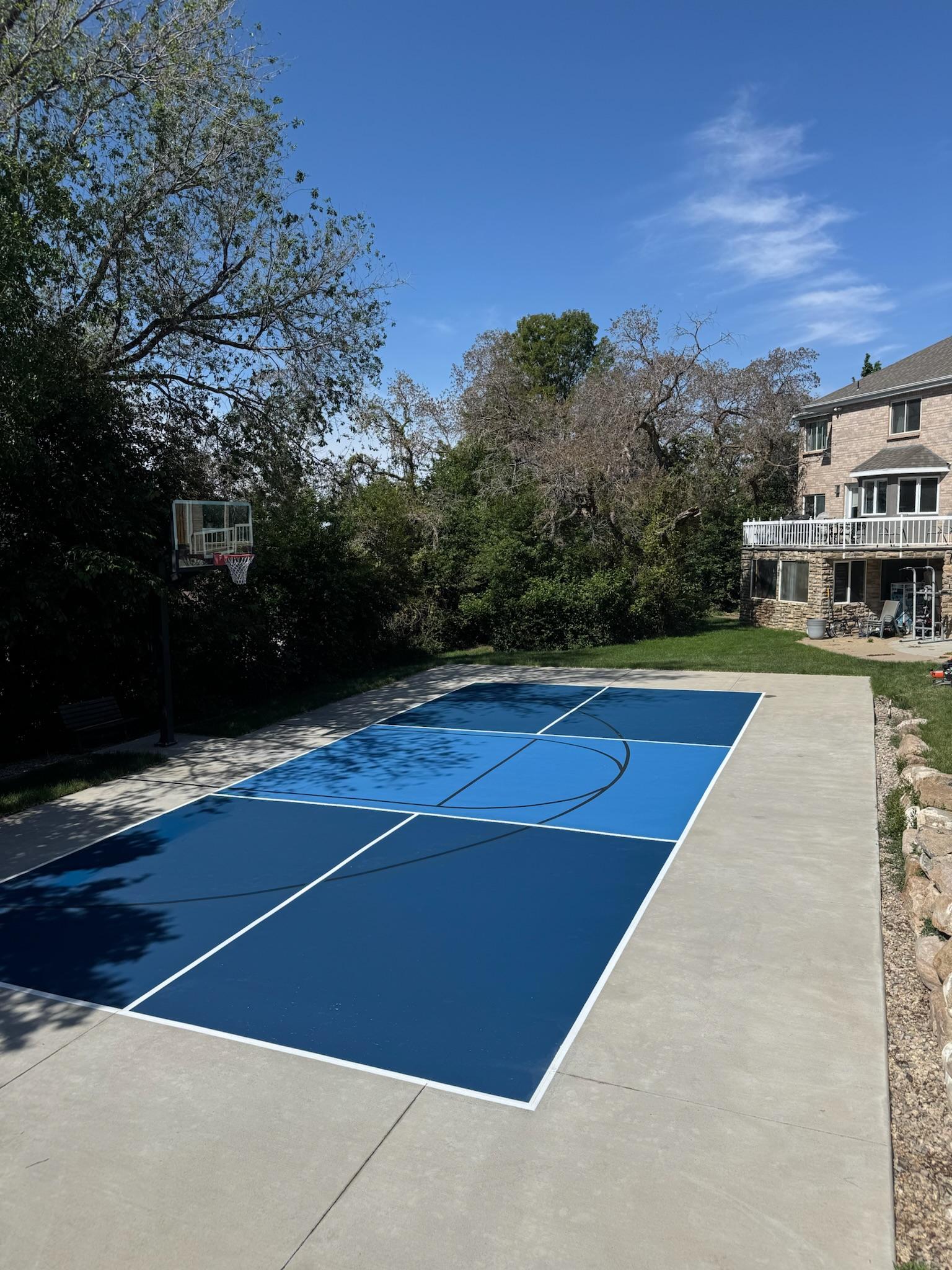 Navy and blue pickleball court with black basketball lines on a large pad beside a brick home